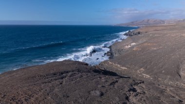 Kuvvetli dalgalarla kayalıklara çarpan kayalık kıyı şeridi berrak bir gökyüzü altında, derin mavi okyanus ve çorak kara arasındaki zıtlığı gözler önüne seriyor..