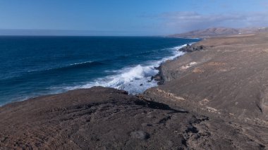 Kuvvetli dalgalarla kayalıklara çarpan kayalık kıyı şeridi berrak bir gökyüzü altında, derin mavi okyanus ve çorak kara arasındaki zıtlığı gözler önüne seriyor..