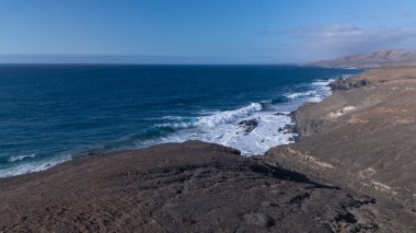 Kuvvetli dalgalarla kayalıklara çarpan kayalık kıyı şeridi berrak bir gökyüzü altında, derin mavi okyanus ve çorak kara arasındaki zıtlığı gözler önüne seriyor..