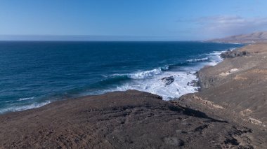 Kuvvetli dalgalarla kayalıklara çarpan kayalık kıyı şeridi berrak bir gökyüzü altında, derin mavi okyanus ve çorak kara arasındaki zıtlığı gözler önüne seriyor..