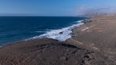 Kuvvetli dalgalarla kayalıklara çarpan kayalık kıyı şeridi berrak bir gökyüzü altında, derin mavi okyanus ve çorak kara arasındaki zıtlığı gözler önüne seriyor..