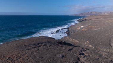 Kuvvetli dalgalarla kayalıklara çarpan kayalık kıyı şeridi berrak bir gökyüzü altında, derin mavi okyanus ve çorak kara arasındaki zıtlığı gözler önüne seriyor..
