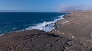 Kuvvetli dalgalarla kayalıklara çarpan kayalık kıyı şeridi berrak bir gökyüzü altında, derin mavi okyanus ve çorak kara arasındaki zıtlığı gözler önüne seriyor..