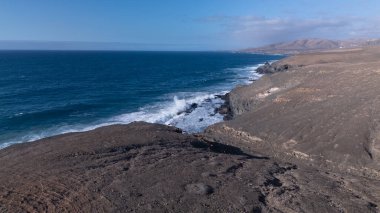 Kuvvetli dalgalarla kayalıklara çarpan kayalık kıyı şeridi berrak bir gökyüzü altında, derin mavi okyanus ve çorak kara arasındaki zıtlığı gözler önüne seriyor..