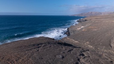 Kuvvetli dalgalarla kayalıklara çarpan kayalık kıyı şeridi berrak bir gökyüzü altında, derin mavi okyanus ve çorak kara arasındaki zıtlığı gözler önüne seriyor..