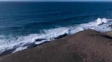 Kuvvetli dalgalarla kayalıklara çarpan kayalık kıyı şeridi berrak bir gökyüzü altında, derin mavi okyanus ve çorak kara arasındaki zıtlığı gözler önüne seriyor..