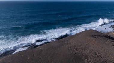 Kuvvetli dalgalarla kayalıklara çarpan kayalık kıyı şeridi berrak bir gökyüzü altında, derin mavi okyanus ve çorak kara arasındaki zıtlığı gözler önüne seriyor..
