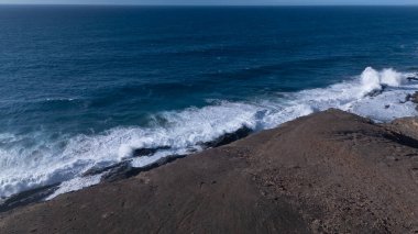 Kuvvetli dalgalarla kayalıklara çarpan kayalık kıyı şeridi berrak bir gökyüzü altında, derin mavi okyanus ve çorak kara arasındaki zıtlığı gözler önüne seriyor..