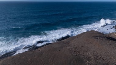 Kuvvetli dalgalarla kayalıklara çarpan kayalık kıyı şeridi berrak bir gökyüzü altında, derin mavi okyanus ve çorak kara arasındaki zıtlığı gözler önüne seriyor..