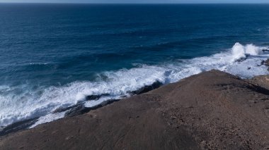 Kuvvetli dalgalarla kayalıklara çarpan kayalık kıyı şeridi berrak bir gökyüzü altında, derin mavi okyanus ve çorak kara arasındaki zıtlığı gözler önüne seriyor..