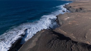 Kuvvetli dalgalarla kayalıklara çarpan kayalık kıyı şeridi berrak bir gökyüzü altında, derin mavi okyanus ve çorak kara arasındaki zıtlığı gözler önüne seriyor..