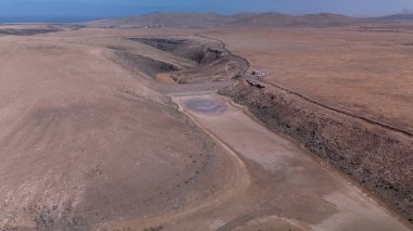 Görüntü, ıssız ve kurak bir arazinin havadan görünüşünü gösteriyor. Olay yerinden geçen toprak bir yol baraja ve neredeyse kurumuş bir su deposuna çıkıyor. Çevredeki arazi kuru olarak karakterize edilir., 