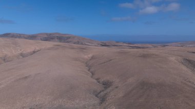 Görüntü, ıssız ve kurak bir arazinin havadan görünüşünü gösteriyor. Olay yerinden geçen toprak bir yol baraja ve neredeyse kurumuş bir su deposuna çıkıyor. Çevredeki arazi kuru olarak karakterize edilir., 