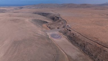 Görüntü, ıssız ve kurak bir arazinin havadan görünüşünü gösteriyor. Olay yerinden geçen toprak bir yol baraja ve neredeyse kurumuş bir su deposuna çıkıyor. Çevredeki arazi kuru olarak karakterize edilir., 