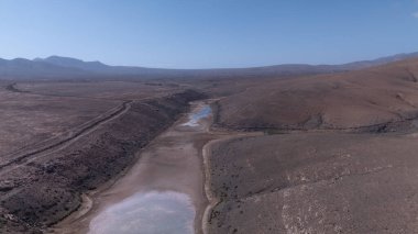 Görüntü, ıssız ve kurak bir arazinin havadan görünüşünü gösteriyor. Olay yerinden geçen toprak bir yol baraja ve neredeyse kurumuş bir su deposuna çıkıyor. Çevredeki arazi kuru olarak karakterize edilir., 