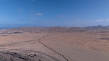 Yuvarlanan kahverengi tepeleri ufka kadar uzanan engin kurak bir manzara. Derin mavi okyanus berrak, parlak bir gökyüzünün altında zıt düşerken bir kanyon kuru araziyi kesiyor..