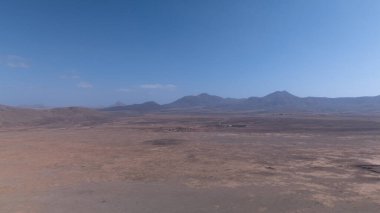 Yuvarlanan kahverengi tepeleri ufka kadar uzanan engin kurak bir manzara. Derin mavi okyanus berrak, parlak bir gökyüzünün altında zıt düşerken bir kanyon kuru araziyi kesiyor..