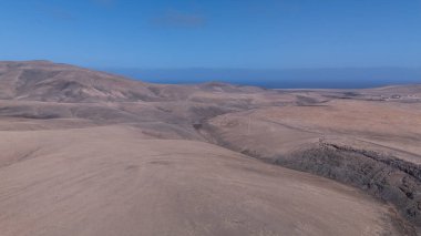 Yuvarlanan kahverengi tepeleri ufka kadar uzanan engin kurak bir manzara. Derin mavi okyanus berrak, parlak bir gökyüzünün altında zıt düşerken bir kanyon kuru araziyi kesiyor..