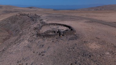 Kuru bir çöl manzarasının havadan görünüşü antik taş kalıntılarını gösteriyor. Daha küçük odalı oval yapı toprak bir yolun yanında duruyor, kayalık araziye karışıyor, sessiz ve gizemli..