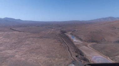 Uçsuz bucaksız, dolambaçlı bir yol, kuru tepeler ve berrak mavi gökyüzünün altındaki uzak dağlar tarafından çerçevelenmiş neredeyse boş bir rezervuar..