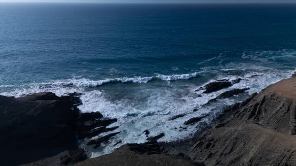  Kuvvetli dalgalarla kayalıklara çarpan kayalık kıyı şeridi berrak bir gökyüzü altında, derin mavi okyanus ve çorak kara arasındaki zıtlığı gözler önüne seriyor..