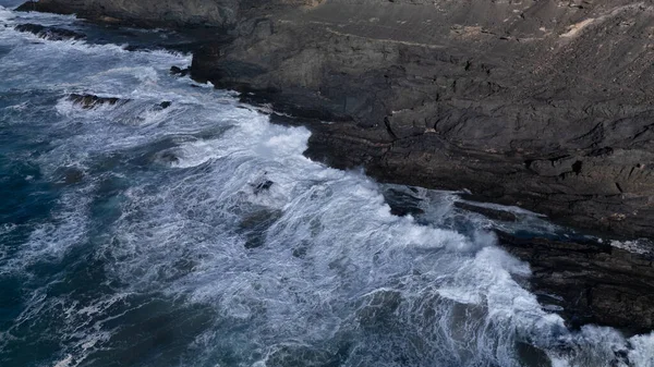 Kuvvetli dalgalarla kayalıklara çarpan kayalık kıyı şeridi berrak bir gökyüzü altında, derin mavi okyanus ve çorak kara arasındaki zıtlığı gözler önüne seriyor..