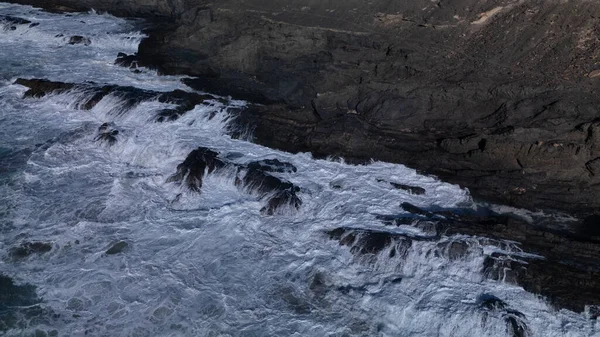 Kuvvetli dalgalarla kayalıklara çarpan kayalık kıyı şeridi berrak bir gökyüzü altında, derin mavi okyanus ve çorak kara arasındaki zıtlığı gözler önüne seriyor..