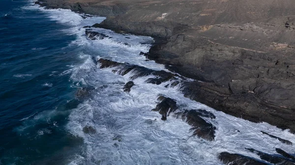 Kuvvetli dalgalarla kayalıklara çarpan kayalık kıyı şeridi berrak bir gökyüzü altında, derin mavi okyanus ve çorak kara arasındaki zıtlığı gözler önüne seriyor..