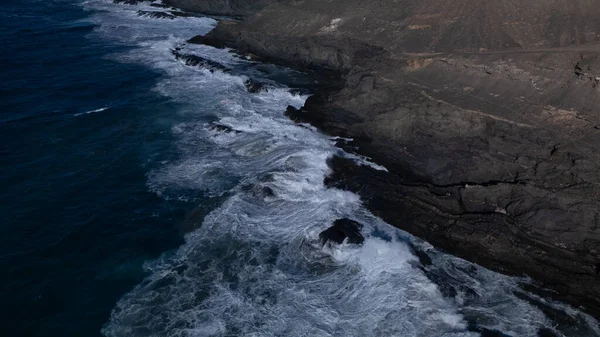 Kuvvetli dalgalarla kayalıklara çarpan kayalık kıyı şeridi berrak bir gökyüzü altında, derin mavi okyanus ve çorak kara arasındaki zıtlığı gözler önüne seriyor..