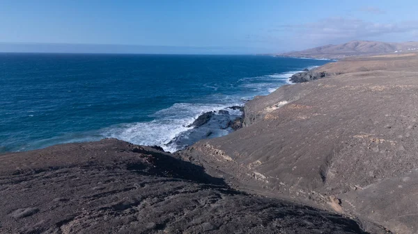 Kuvvetli dalgalarla kayalıklara çarpan kayalık kıyı şeridi berrak bir gökyüzü altında, derin mavi okyanus ve çorak kara arasındaki zıtlığı gözler önüne seriyor..