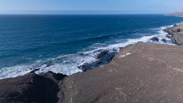 Kuvvetli dalgalarla kayalıklara çarpan kayalık kıyı şeridi berrak bir gökyüzü altında, derin mavi okyanus ve çorak kara arasındaki zıtlığı gözler önüne seriyor..