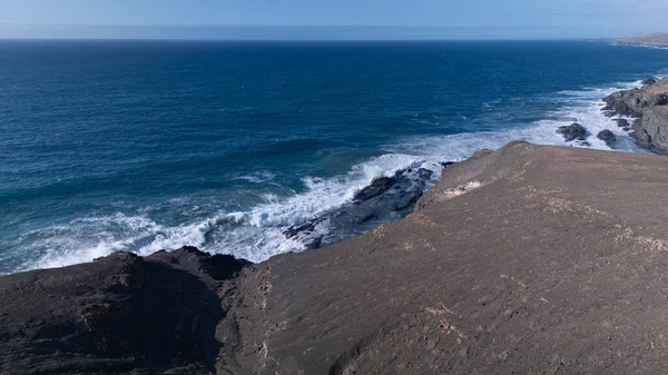 Kuvvetli dalgalarla kayalıklara çarpan kayalık kıyı şeridi berrak bir gökyüzü altında, derin mavi okyanus ve çorak kara arasındaki zıtlığı gözler önüne seriyor..