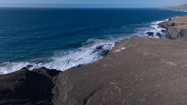 Kuvvetli dalgalarla kayalıklara çarpan kayalık kıyı şeridi berrak bir gökyüzü altında, derin mavi okyanus ve çorak kara arasındaki zıtlığı gözler önüne seriyor..