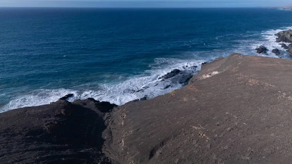 Kuvvetli dalgalarla kayalıklara çarpan kayalık kıyı şeridi berrak bir gökyüzü altında, derin mavi okyanus ve çorak kara arasındaki zıtlığı gözler önüne seriyor..