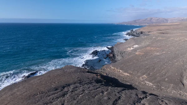Kuvvetli dalgalarla kayalıklara çarpan kayalık kıyı şeridi berrak bir gökyüzü altında, derin mavi okyanus ve çorak kara arasındaki zıtlığı gözler önüne seriyor..