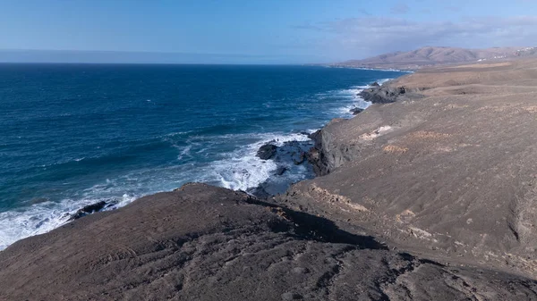 Kuvvetli dalgalarla kayalıklara çarpan kayalık kıyı şeridi berrak bir gökyüzü altında, derin mavi okyanus ve çorak kara arasındaki zıtlığı gözler önüne seriyor..