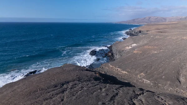 Kuvvetli dalgalarla kayalıklara çarpan kayalık kıyı şeridi berrak bir gökyüzü altında, derin mavi okyanus ve çorak kara arasındaki zıtlığı gözler önüne seriyor..