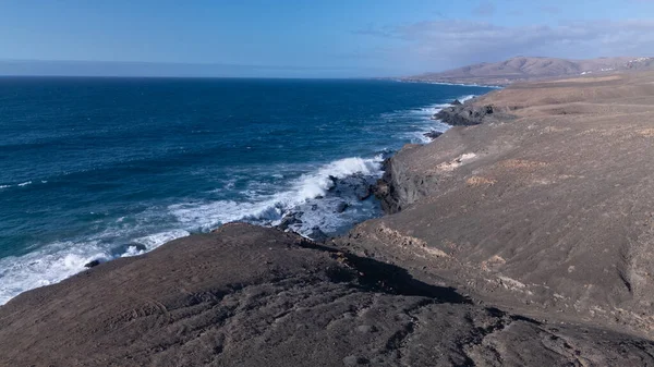 Kuvvetli dalgalarla kayalıklara çarpan kayalık kıyı şeridi berrak bir gökyüzü altında, derin mavi okyanus ve çorak kara arasındaki zıtlığı gözler önüne seriyor..