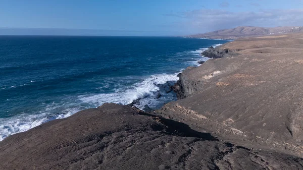 Kuvvetli dalgalarla kayalıklara çarpan kayalık kıyı şeridi berrak bir gökyüzü altında, derin mavi okyanus ve çorak kara arasındaki zıtlığı gözler önüne seriyor..