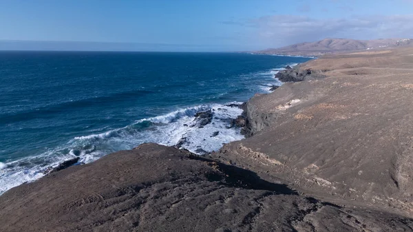 Kuvvetli dalgalarla kayalıklara çarpan kayalık kıyı şeridi berrak bir gökyüzü altında, derin mavi okyanus ve çorak kara arasındaki zıtlığı gözler önüne seriyor..