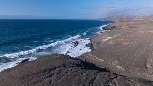 Kuvvetli dalgalarla kayalıklara çarpan kayalık kıyı şeridi berrak bir gökyüzü altında, derin mavi okyanus ve çorak kara arasındaki zıtlığı gözler önüne seriyor..