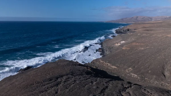 Kuvvetli dalgalarla kayalıklara çarpan kayalık kıyı şeridi berrak bir gökyüzü altında, derin mavi okyanus ve çorak kara arasındaki zıtlığı gözler önüne seriyor..