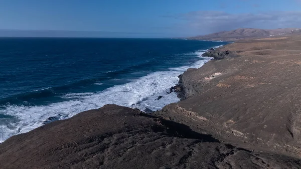 Kuvvetli dalgalarla kayalıklara çarpan kayalık kıyı şeridi berrak bir gökyüzü altında, derin mavi okyanus ve çorak kara arasındaki zıtlığı gözler önüne seriyor..