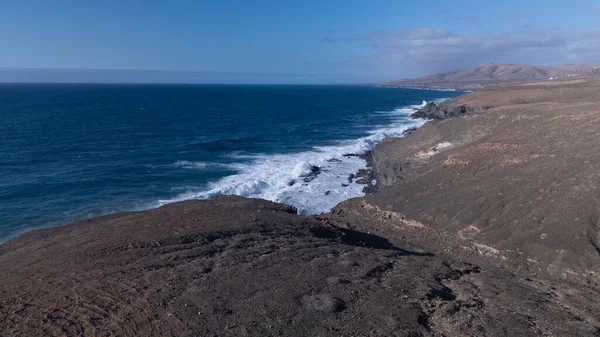 Kuvvetli dalgalarla kayalıklara çarpan kayalık kıyı şeridi berrak bir gökyüzü altında, derin mavi okyanus ve çorak kara arasındaki zıtlığı gözler önüne seriyor..
