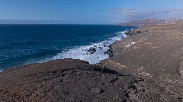 Kuvvetli dalgalarla kayalıklara çarpan kayalık kıyı şeridi berrak bir gökyüzü altında, derin mavi okyanus ve çorak kara arasındaki zıtlığı gözler önüne seriyor..