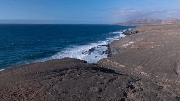 Kuvvetli dalgalarla kayalıklara çarpan kayalık kıyı şeridi berrak bir gökyüzü altında, derin mavi okyanus ve çorak kara arasındaki zıtlığı gözler önüne seriyor..