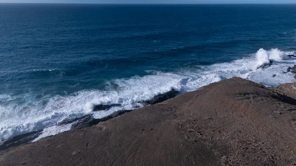 Kuvvetli dalgalarla kayalıklara çarpan kayalık kıyı şeridi berrak bir gökyüzü altında, derin mavi okyanus ve çorak kara arasındaki zıtlığı gözler önüne seriyor..