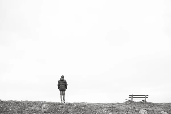 Una sola persona de pie junto a un banco de madera vacío en el horizonte — Foto de Stock