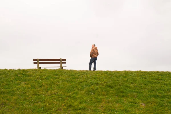 A single person standing next to a single empty wooden bench on the horizon