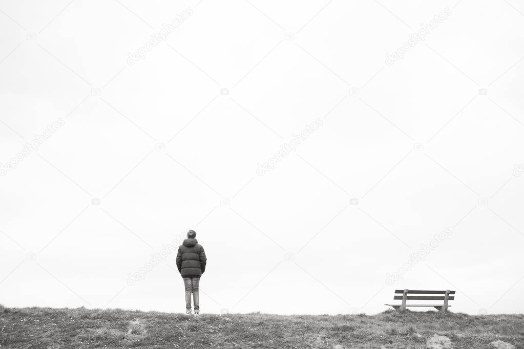 A single person standing next to a single empty wooden bench on the horizon